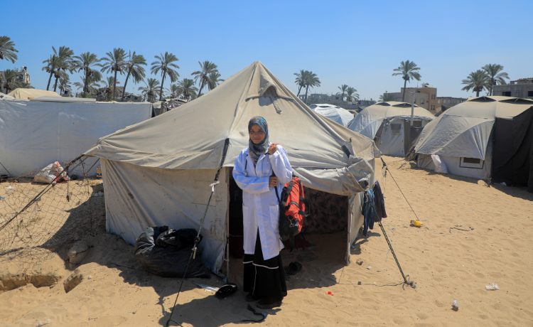 A woman in a white lab coat stands outside a tent in a displacement camp